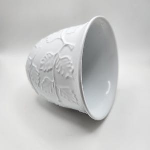 May include: White ceramic planter with embossed leaf and vine design. The planter has a slightly flared rim and a smooth interior. The exterior features a repeating pattern of leaves and vines. The planter is suitable for small plants or decorative use.