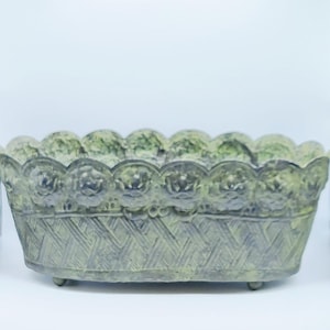 May include: An oval-shaped, antique-style metal planter with a distressed green and gray finish. The rim features a scalloped edge with floral detailing, and the body has a woven basket design. The planter rests on small, rounded feet.