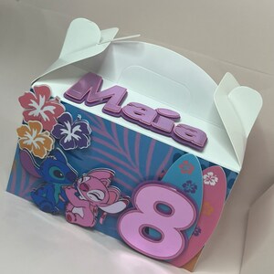 May include: A white party favor box with a blue and pink tropical design. The box features the name "Maia" in purple letters, the number "8", and images of Stitch and Angel. Decorative flowers and surfboards are also included.
