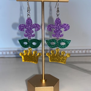 Puede incluir: Un par de pendientes colgantes con un lirio morado, una máscara verde y un dije de corona dorada. Los pendientes están en un soporte dorado.