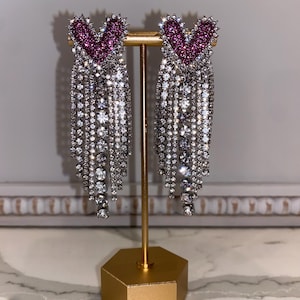 May include: A pair of long, dangling earrings with a pink heart-shaped charm at the top and a cascade of clear rhinestones.