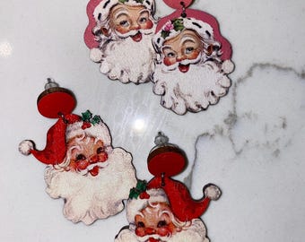 May include: Wooden Santa Claus earrings. The earrings feature a vintage-style Santa face with a red hat, white beard, and rosy cheeks. The earrings are attached to a pink wooden circle. Festive holiday jewellery.
