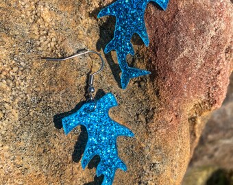 May include: A pair of blue glitter hammerhead shark earrings. The earrings are made of a shiny, glittery material and have silver-colored fish hook ear wires. The earrings are displayed on a textured, brown rock surface.
