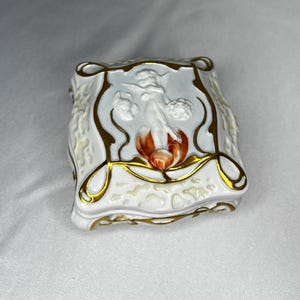 Puede incluir: Una caja de porcelana blanca con un diseño floral dorado y naranja y una imagen en relieve de un querubín. La caja tiene forma cuadrada con esquinas redondeadas y una tapa decorativa.