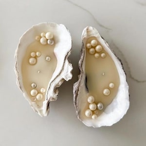 May include: Two oyster shell candles with a creamy wax interior and embedded pearl-like beads. The shells are white with gray and brown edges, resting on a light-colored surface. The candles have a coastal or nautical theme.