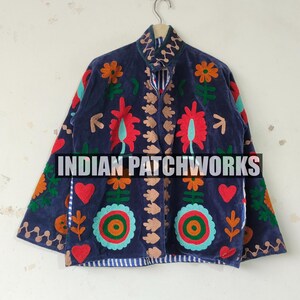 Pode incluir: Um casaco azul-escuro com bordados vibrantes de flores e corações em várias cores. A peça tem gola alta, mangas compridas e um forro às riscas azuis e brancas visível nas extremidades. O texto INDIAN PATCHWORKS está centralizado.