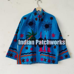 May include: A blue jacket with a vibrant design featuring palm trees, sunbursts, and geometric patterns in red, orange, and black. The jacket has a collared neckline and is hanging on a wooden hanger. The text "Indian Patchworks" is visible.