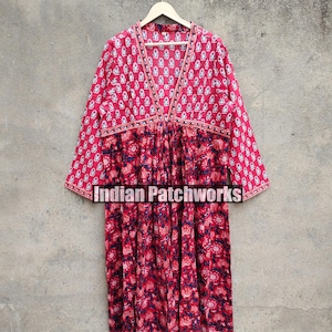 Può includere: Un abito in cotone con stampa floreale rosso e blu, scollo a V e maniche lunghe. L'abito ha una cintura da annodare in vita e una gonna plissettata. L'abito è appeso a un appendiabiti di legno contro un muro grigio. "Indian Patchworks" è stampato sull'abito.
