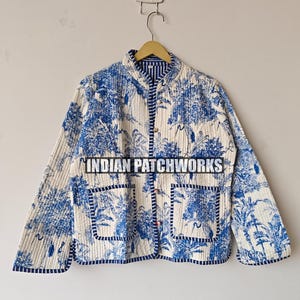 May include: A quilted jacket featuring a blue and white floral design. The jacket has a mandarin collar, two front pockets, and a button-up closure. The trim and lining are a dark blue striped fabric. The text "INDIAN PATCHWORKS" is visible.