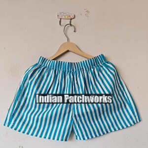 May include: Blue and white striped shorts hanging on a wooden hanger. The shorts have vertical stripes and the text "Indian Patchworks" across the front. The shorts are made of a lightweight fabric.