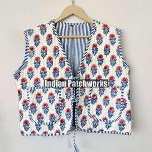 May include: White quilted vest with a floral print in shades of pink and blue. The vest has a V-neckline and a tie closure. It features two pockets on the front and a blue and white striped lining.