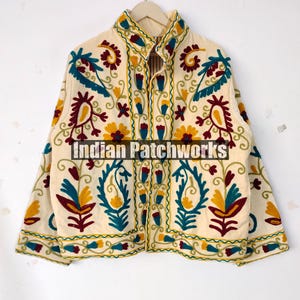 May include: Cream-colored jacket with intricate embroidered floral patterns in yellow, teal, and burgundy. The jacket features a high collar and is suspended from a hanger. The text "Indian Patchworks" is visible on the jacket.