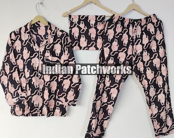 Peut inclure: Un ensemble de pyjama noir avec un motif de chat rose. L'ensemble comprend une chemise boutonnée, un pantalon et un masque pour les yeux assorti. La chemise et le pantalon ont une bordure blanche. L'ensemble de pyjama est suspendu à des cintres en bois.