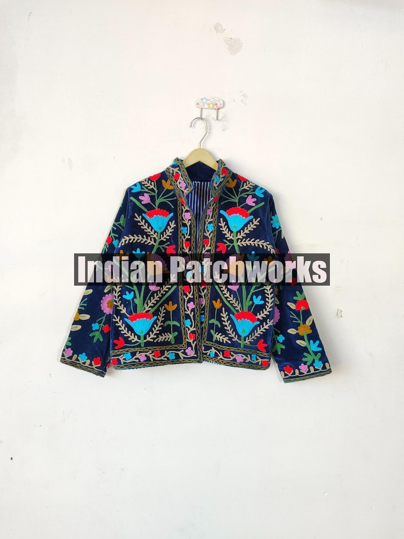 May include: A navy blue velvet jacket with colorful floral embroidery. The jacket has a mandarin collar and long sleeves. The embroidery features a variety of flowers, leaves, and vines in shades of red, pink, yellow, blue, and green. The text "Indian Patchworks" is visible on the jacket.
