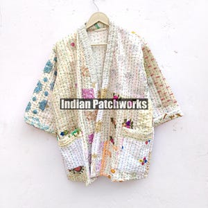 K&ouml;nnte beinhalten: Eine wei&szlig;e gesteppte Jacke mit Patchwork-Muster und bunten Stickereien. Die Jacke hat einen Kragen und einen vorderen &Ouml;ffnung. Die Jacke h&auml;ngt an einem Holzb&uuml;gel an einer wei&szlig;en Wand. Der Text "Indian Patchworks" ist auf der Jacke sichtbar.