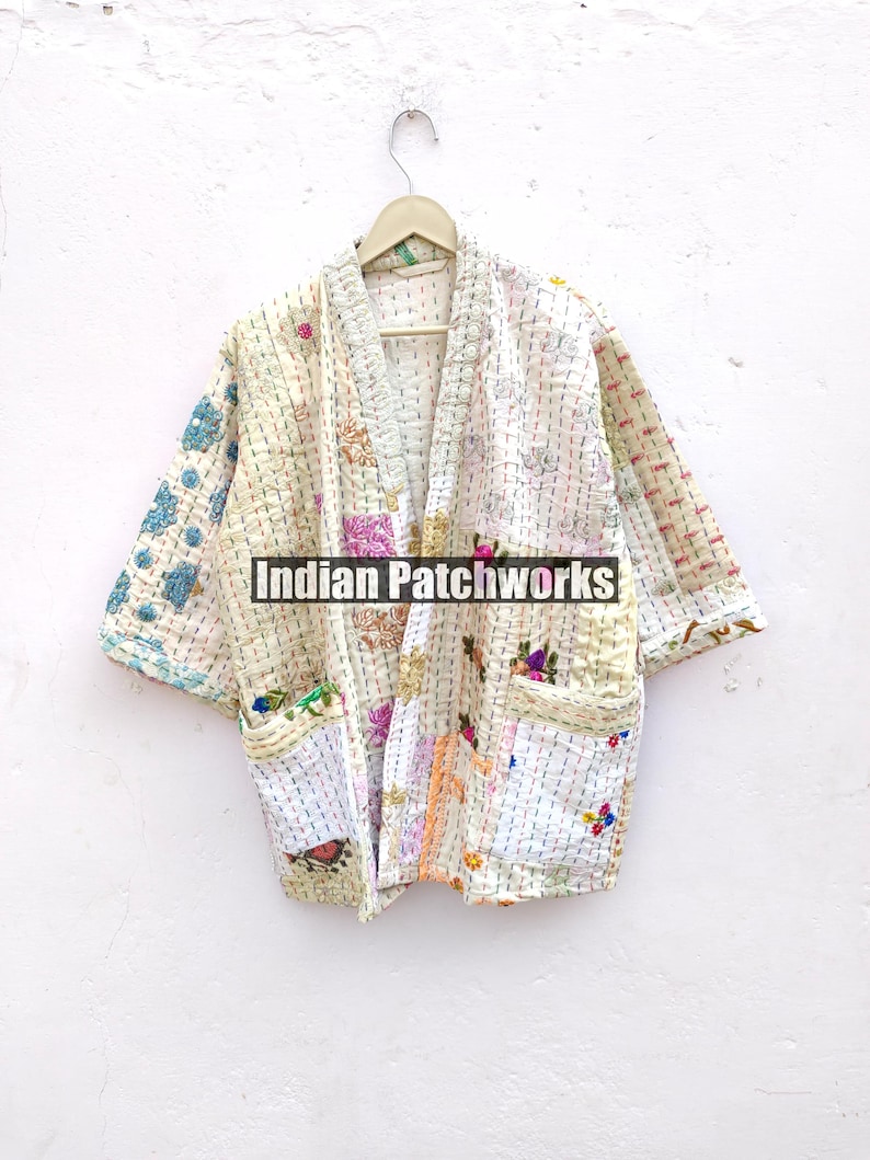 K&ouml;nnte beinhalten: Eine wei&szlig;e gesteppte Jacke mit Patchwork-Muster und bunten Stickereien. Die Jacke hat einen Kragen und einen vorderen &Ouml;ffnung. Die Jacke h&auml;ngt an einem Holzb&uuml;gel an einer wei&szlig;en Wand. Der Text "Indian Patchworks" ist auf der Jacke sichtbar.