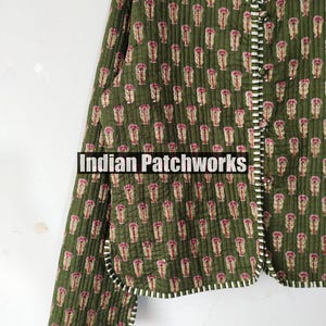 May include: Olive green quilted jacket with a repeating pink floral pattern. The jacket has a white and black striped trim and the text "Indian Patchworks". The jacket is buttoned up and has long sleeves.