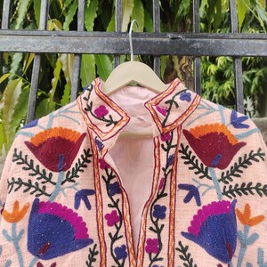 May include: A pink embroidered jacket with a floral design. The embroidery features a variety of colours, including orange, red, blue, and green.