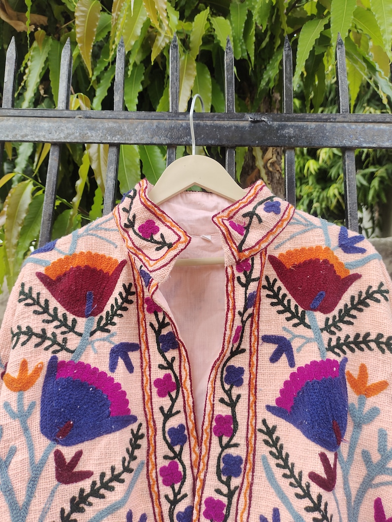 May include: A pink embroidered jacket with a floral design. The embroidery features a variety of colours, including orange, red, blue, and green.