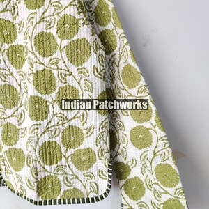 May include: A quilted jacket with a floral design. The jacket has a white base, adorned with a repeating pattern of green flowers and vines. The edges are finished with a black and white striped trim. The text "Indian Patchworks" is visible.