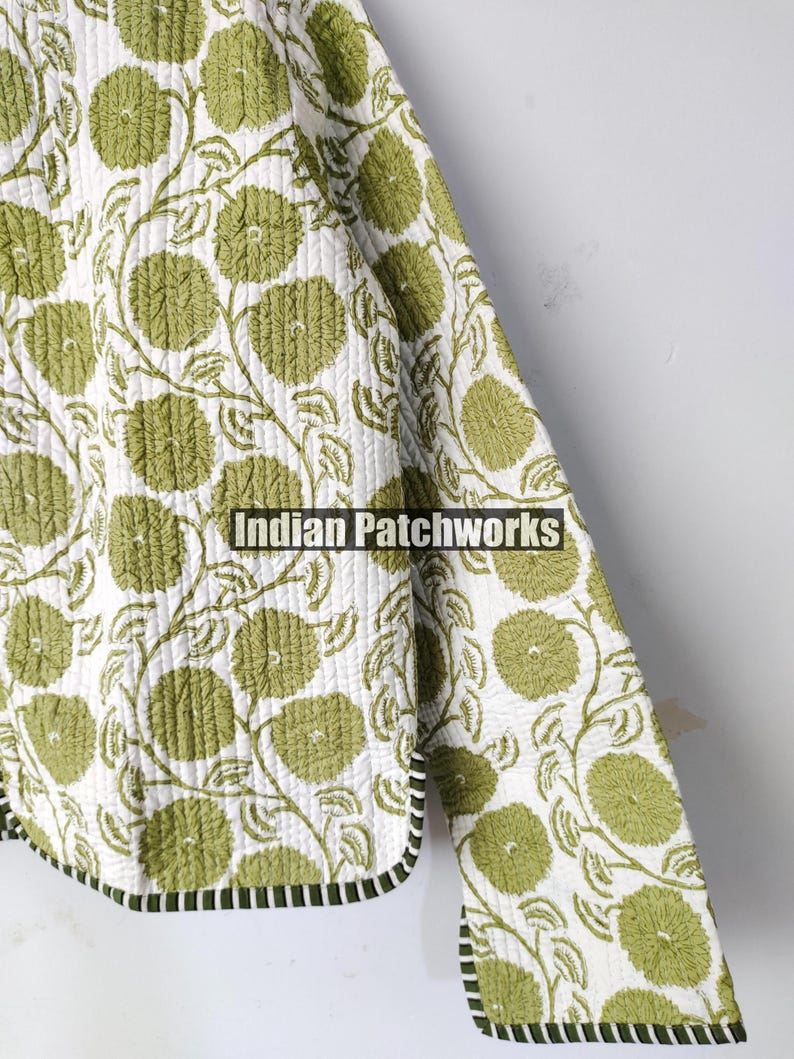 May include: A quilted jacket with a floral design. The jacket has a white base, adorned with a repeating pattern of green flowers and vines. The edges are finished with a black and white striped trim. The text "Indian Patchworks" is visible.