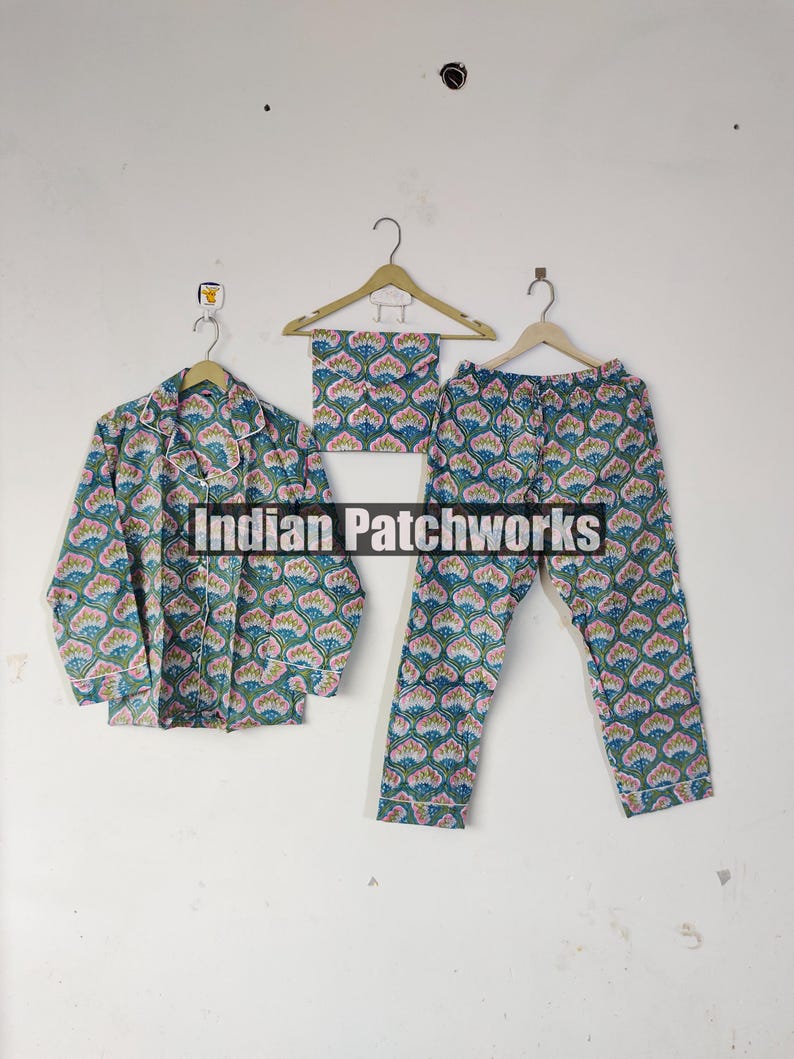 May include: A three-piece pajama set featuring a button-down shirt, pants, and a matching eye mask. The set is adorned with a repeating floral pattern in shades of teal, pink, and white. The text "Indian Patchworks" is visible.