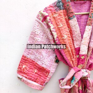 May include: A pink and peach patchwork kimono robe with a floral print and embroidered details. The robe has a tie belt and is made of quilted fabric. "Indian Patchworks" is printed on the robe.