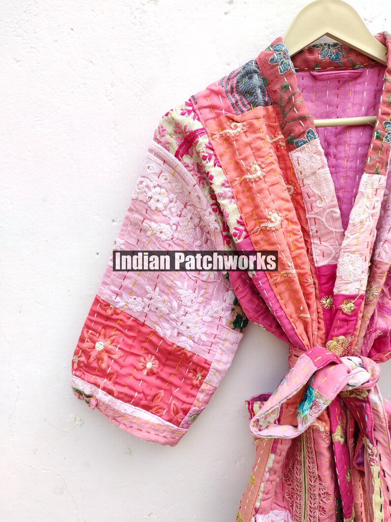 May include: A pink and peach patchwork kimono robe with a floral print and embroidered details. The robe has a tie belt and is made of quilted fabric. "Indian Patchworks" is printed on the robe.
