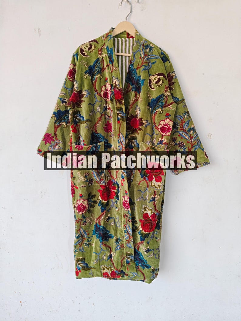 May include: A green velvet robe with a floral print. The robe has a long, flowing silhouette and a wide collar. The robe is open at the front, revealing a white and green striped lining. The robe has two pockets on the front. The text "Indian Patchworks" is visible on the robe.