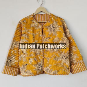 Könnte beinhalten: Eine gelbe gesteppte Jacke mit Blumendruck und gestreiftem Besatz. Die Jacke hat einen Knopfverschluss und lange Ärmel. Der Text "Indian Patchworks" ist auf der Vorderseite der Jacke aufgedruckt.