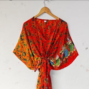 May include: A vibrant, flowing, red and orange patterned kaftan dress with a V-neck and tie closure. The fabric features a floral design and a colorful illustration of figures. The dress is hanging on a wooden hanger.