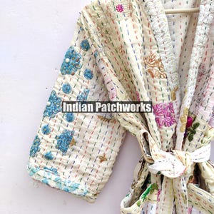 K&ouml;nnte beinhalten: Eine wei&szlig;e Baumwollrobe mit bunten Stickereien und einem Bindeg&uuml;rtel. Die Robe ist aus Patchwork-Stoff gefertigt und hat einen Vintage-Look.