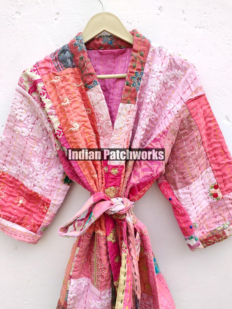 May include: A pink and peach patchwork kimono robe with a tie belt. The robe is made of quilted fabric and has a floral design. The text "Indian Patchworks" is printed on the robe.