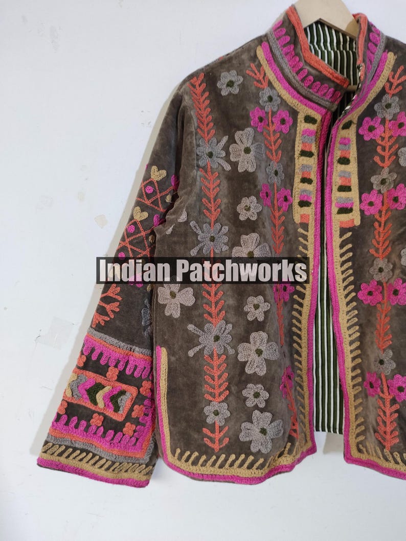 May include: A brown embroidered jacket with floral and geometric patterns. The jacket features pink, grey, and orange accents, with the text "Indian Patchworks" visible. The jacket has a striped lining and a high collar.