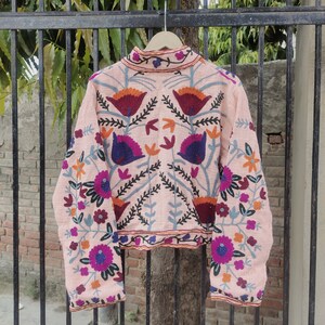 May include: A pink embroidered jacket with floral patterns in shades of blue, purple, orange, and red. The jacket has long sleeves and a high neckline.