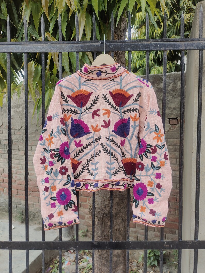 May include: A pink embroidered jacket with floral patterns in shades of blue, purple, orange, and red. The jacket has long sleeves and a high neckline.