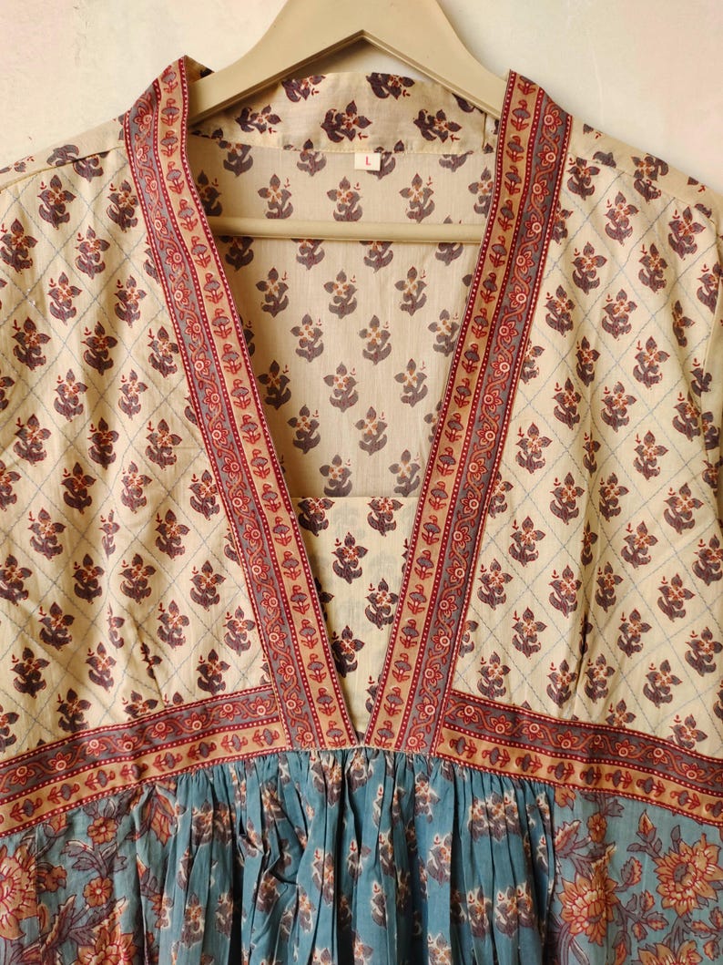 May include: A cream-colored cotton dress with a V-neckline and a blue floral print. The dress has a red and cream trim around the neckline and sleeves. The dress is hanging on a wooden hanger.