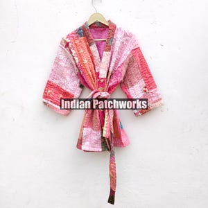 May include: A pink and red patchwork quilted kimono with a tie belt. The kimono is hanging on a white hanger against a white wall. The text "Indian Patchworks" is visible in the image.