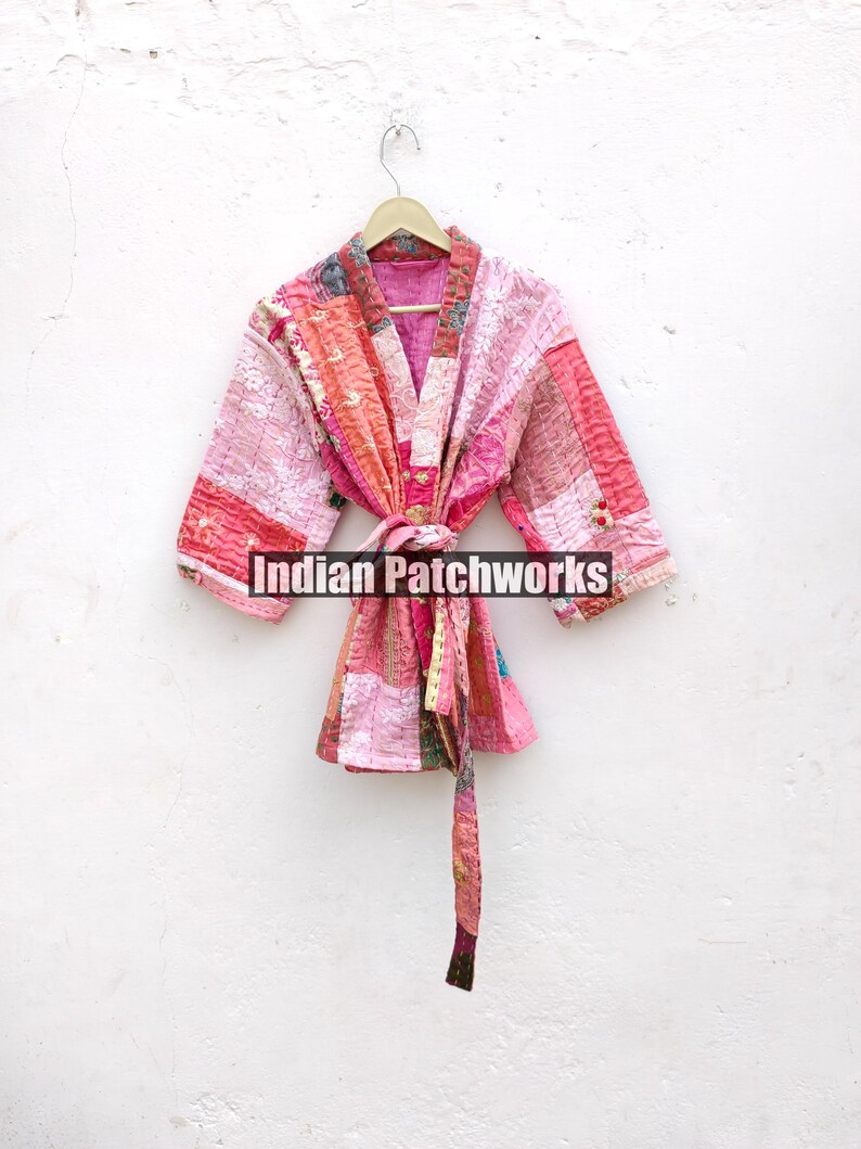 May include: A pink and red patchwork quilted kimono with a tie belt. The kimono is hanging on a white hanger against a white wall. The text "Indian Patchworks" is visible in the image.