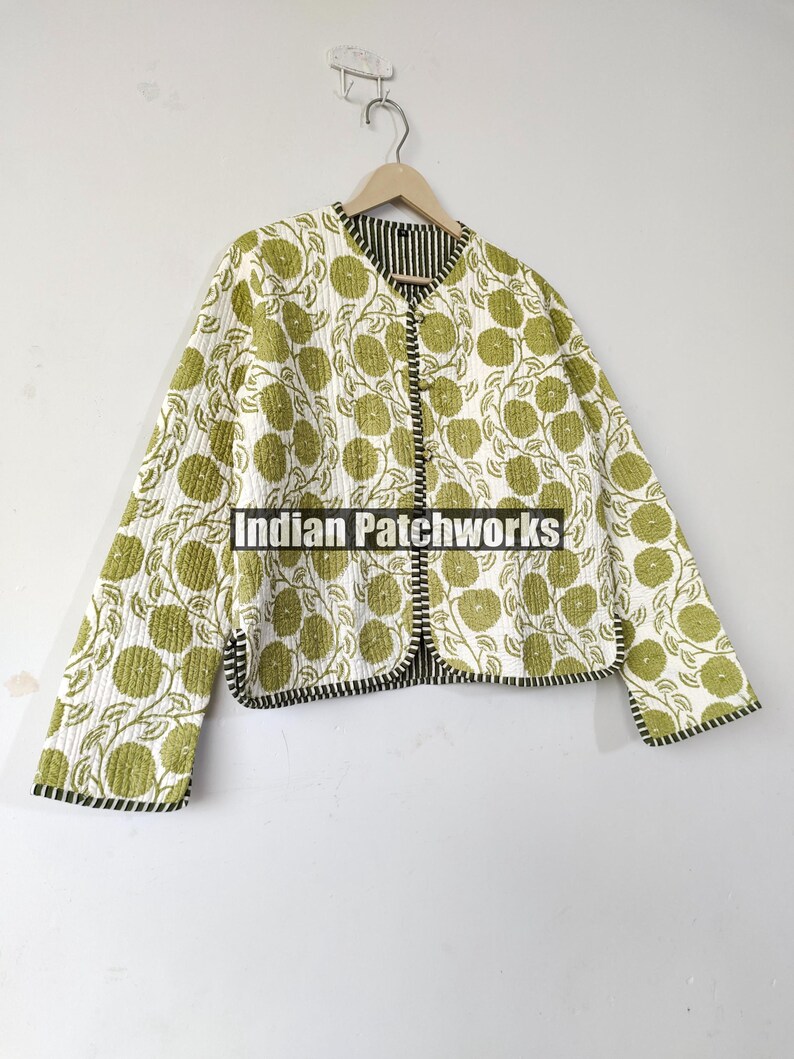 May include: A quilted jacket with a white background and a repeating pattern of green floral designs. The jacket has a black and white striped lining and trim. The jacket is hanging on a wooden hanger.