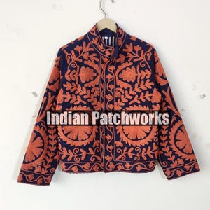 Op de afbeelding: Marineblauwe jas met ingewikkelde oranje bloemenborduurwerk. De jas heeft een hoge kraag en lange mouwen. De tekst "Indian Patchworks" staat op de voorkant. De jas hangt aan een metalen hanger.