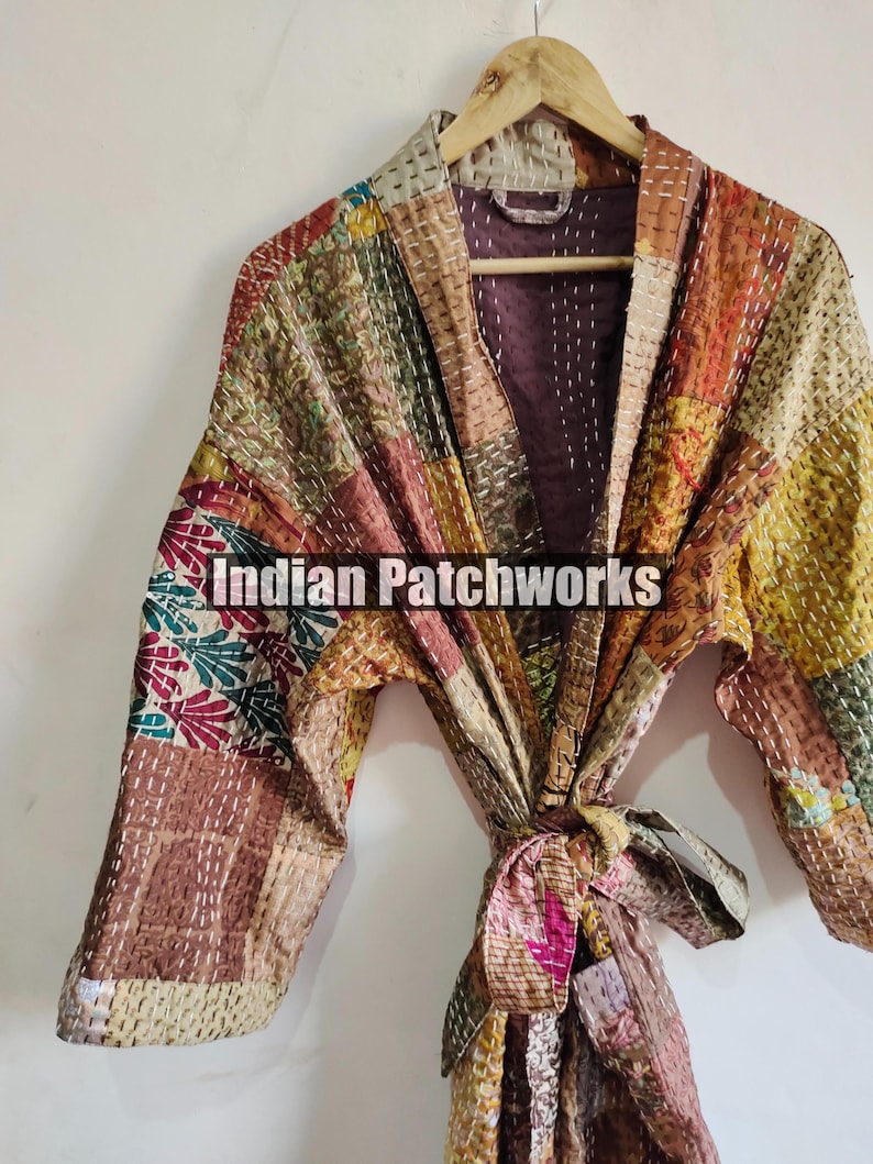 May include: A patchwork quilted robe with a tie belt. The robe is made of various fabrics in shades of brown, beige, pink, and green. The fabric is stitched together in a patchwork pattern. The robe has a long sleeve and a V-neck. The robe is hanging on a wooden hanger. The text "Indian Patchworks" is visible on the robe.