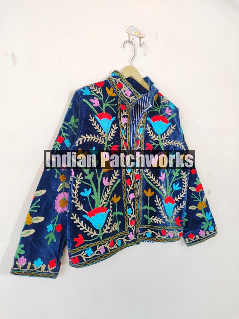 May include: A navy blue velvet jacket with colorful floral embroidery. The jacket has a mandarin collar and a front zipper closure. The embroidery is intricate and detailed, featuring a variety of flowers and leaves in shades of red, pink, yellow, blue, and green. The jacket is lined with a striped fabric.