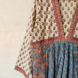May include: A long-sleeved, cream-colored dress with a V-neckline and a blue and orange floral print. The dress has a gathered skirt and a red and white trim around the neckline and sleeves.