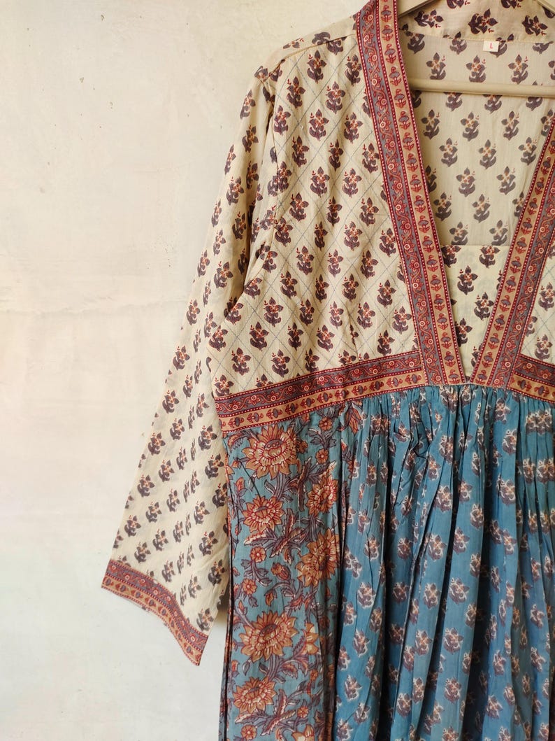 May include: A long-sleeved, cream-colored dress with a V-neckline and a blue and orange floral print. The dress has a gathered skirt and a red and white trim around the neckline and sleeves.