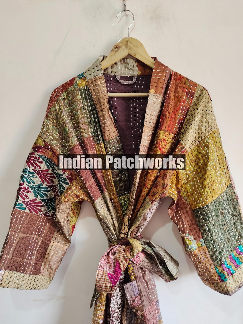 May include: A patchwork quilted robe with a tie belt. The robe is made of various fabrics in shades of brown, green, and orange. The fabric is stitched together in a patchwork pattern. The robe has a long sleeve and a collar. The robe is tied at the waist with a matching tie belt. The text "Indian Patchworks" is printed on the robe.