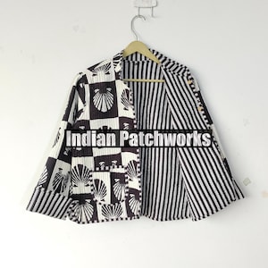 Op de afbeelding: Een zwart-wit gewatteerde jas met een schelpenpatroon op de voorkant en een gestreepte binnenkant. De jas hangt aan een houten hanger. De tekst "Indian Patchworks" is zichtbaar op de voorkant.