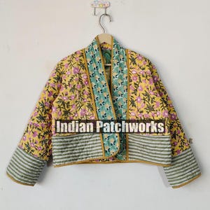 Peut inclure: Une veste matelassée à motifs floraux jaunes, verts et roses avec une bordure rayée. La veste est ouverte à l'avant et a un style long et fluide. Le texte "Indian Patchworks" est imprimé sur le devant de la veste.
