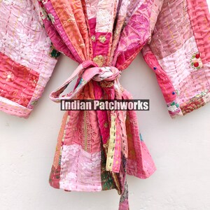 May include: A pink and white patchwork kimono with a long sash. The kimono is made of different fabrics and has a quilted texture. The sash is tied at the waist and has a tassel at the end. The kimono is embroidered with floral designs. The text "Indian Patchworks" is visible on the sash.