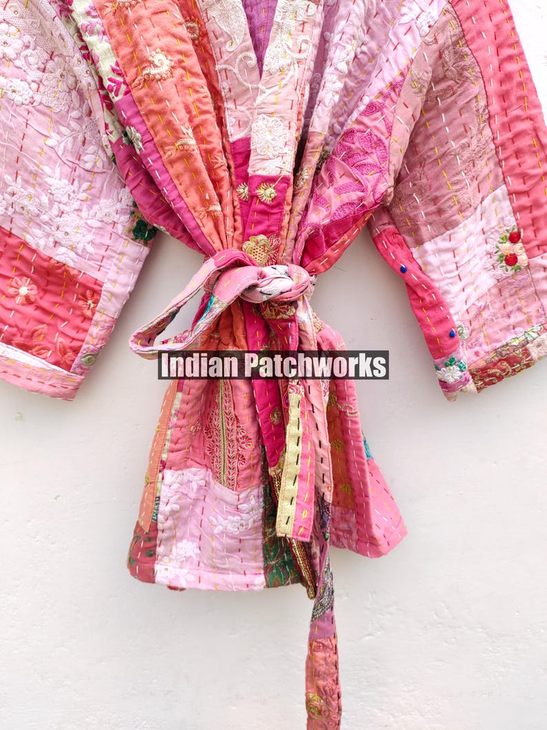 May include: A pink and white patchwork kimono with a long sash. The kimono is made of different fabrics and has a quilted texture. The sash is tied at the waist and has a tassel at the end. The kimono is embroidered with floral designs. The text "Indian Patchworks" is visible on the sash.